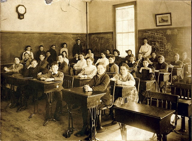 Sepia-toned historical photo of a classroom with over twenty students, seated at individual wooden desks, and a female teacher standing by the blackboard.