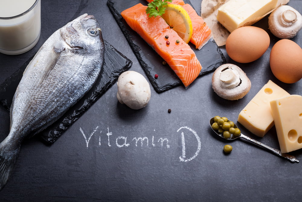 Overhead flat lay of Vitamin D rich foods including fish, milk, eggs, cheese, and mushrooms, with "Vitamin D" written in chalk.
