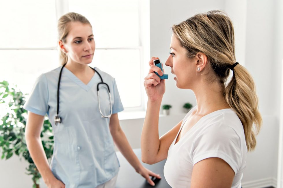 A female patient with blonde hair tied back is using a blue inhaler likely an asthma rescue inhaler by holding it up to her mouth and inhaling.