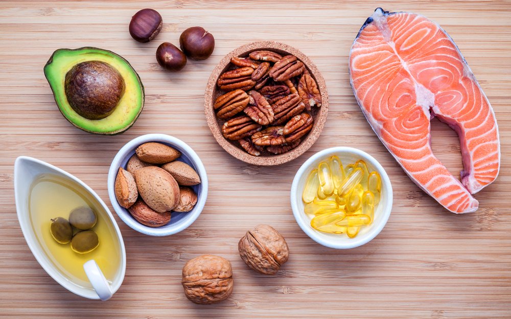 Overhead flat lay of healthy fat sources including salmon, avocado, pecans, almonds, walnuts, olive oil, and Omega-3 supplement capsules.