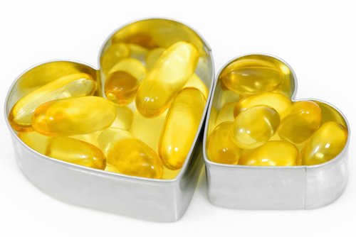 A collection of shiny, translucent gold soft gel capsules likely fish oil or omega-3 supplements spilling out of two heart-shaped cookie cutters. The image emphasizes the heart health benefits of the supplements. The background is solid white.