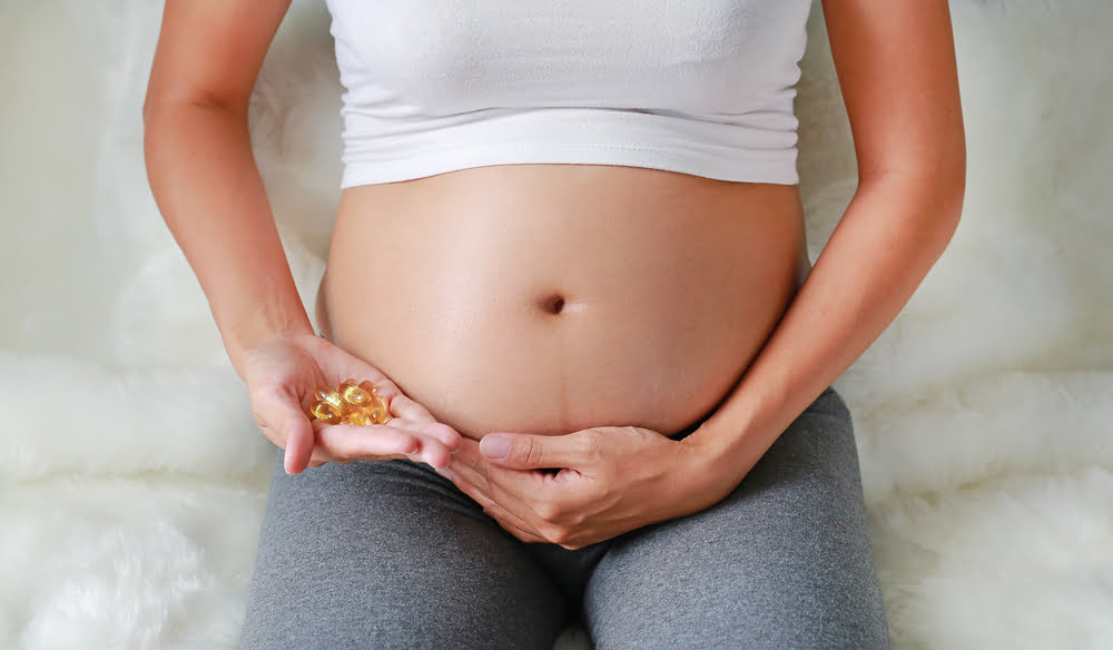 A close-up view of a pregnant woman's bare abdomen. She is wearing a white crop top and gray leggings, sitting on a white fuzzy surface.