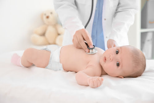 A close-up of a baby lying on a white examination table, wearing only a diaper and pink socks.