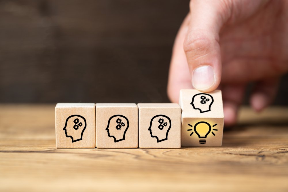 A hand is placing a wooden block in a row of four on a wooden surface. Three of the blocks show an icon of a human head with gear cogs inside, representing knowledge or process.