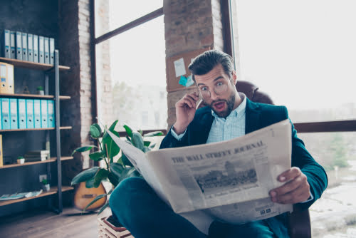 A surprised businessman in a suit reading The Wall Street Journal newspaper in a modern office.