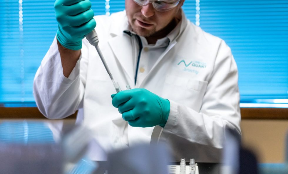 A scientist wearing a white lab coat, protective glasses, and teal gloves is focused on transferring a liquid using a micropipette into a small test tube or vial.