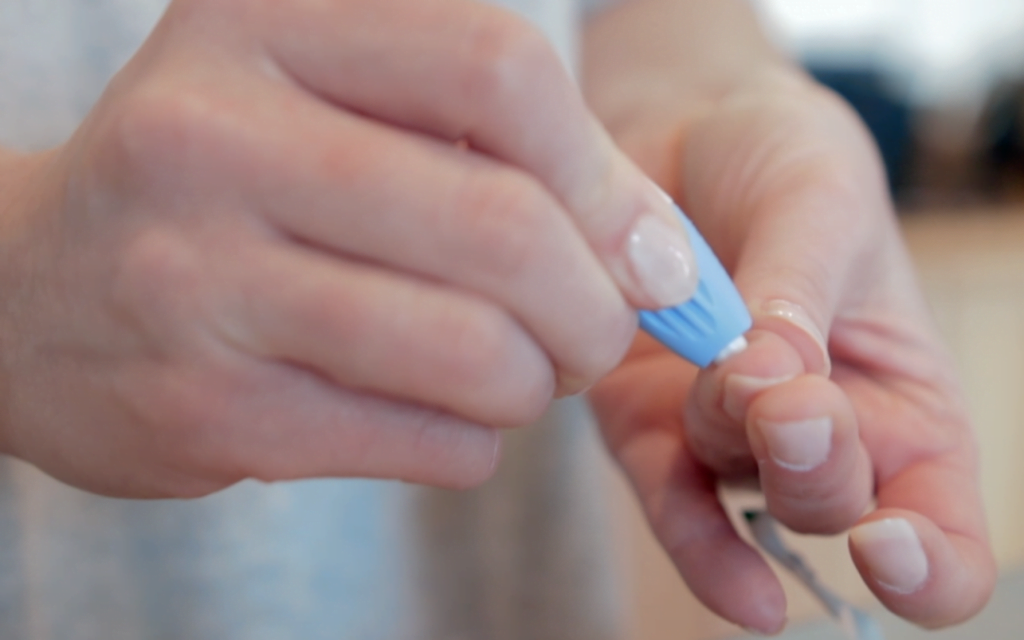 A close-up shot of a person's hands performing a finger-prick test, likely for blood sugar or a small blood sample collection.