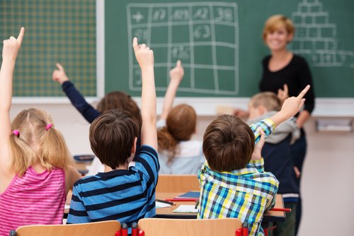 group of elementary school children sitting at desks in a classroom with their backs to the camera, all enthusiastically raising their hands to answer a question.