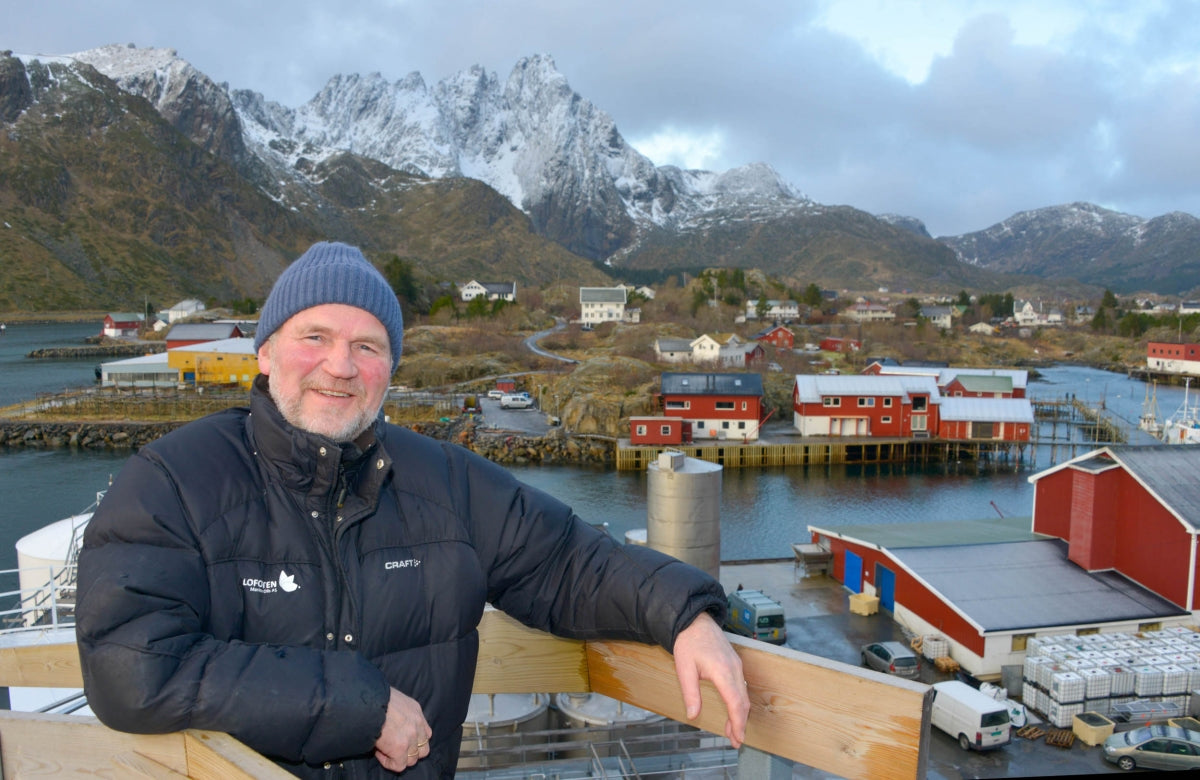A smiling, middle-aged man wearing a dark blue beanie and a heavy black puffer jacket stands on an observation deck with a wooden railing.