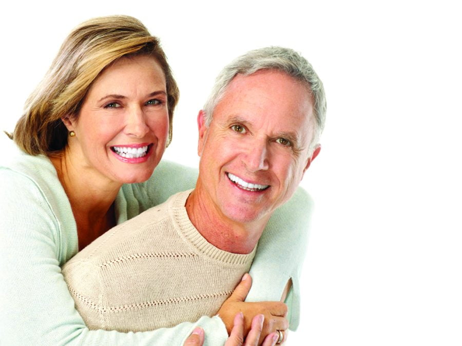 A smiling, middle-aged or senior couple embracing each other against a bright white background. The woman, with blonde hair, is standing behind the man, who has gray hair, and has her arms wrapped around him.