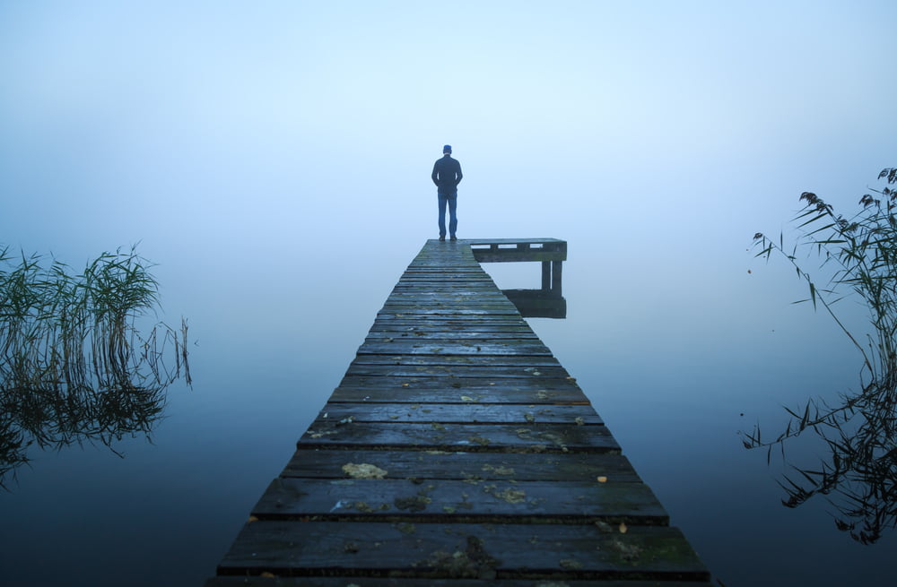 A solitary figure, wearing a baseball cap and jacket, stands at the end of a long, dark, wooden jetty or dock that extends straight out over still, dark water.