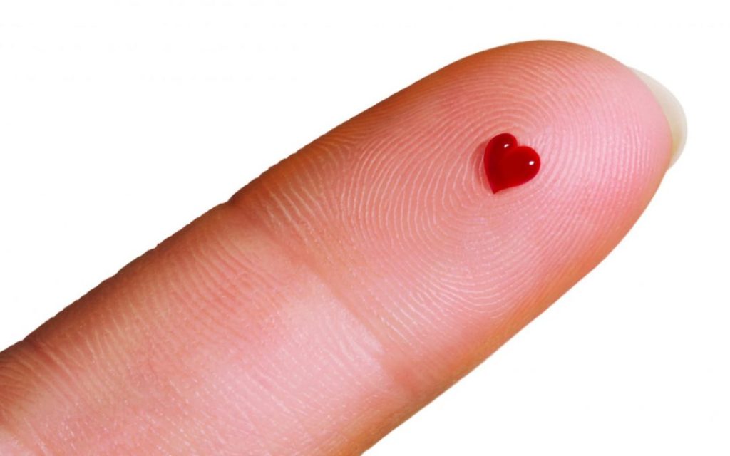 A single human finger with a tiny drop of blood on the fingertip that is perfectly shaped like a small red heart.
