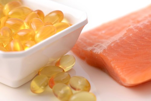 Close-up of omega-3 fish oil capsules in a white bowl next to a raw salmon fillet on a white surface.