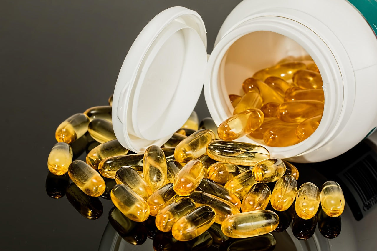 A white plastic bottle of supplements is tipped on its side, with the lid open. A large pile of translucent, golden-yellow soft gel capsules (likely fish oil or other vitamin supplements) has spilled out onto a dark, reflective surface.