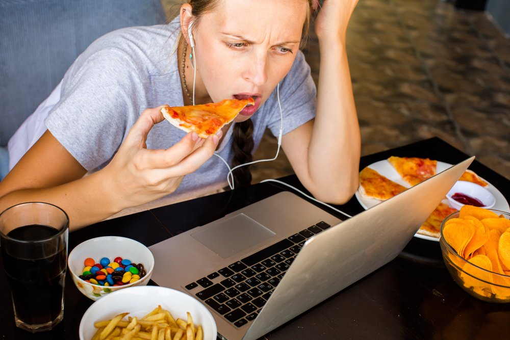 A focused woman, wearing a gray t-shirt and earbuds, is working on a laptop while simultaneously eating a slice of pizza.