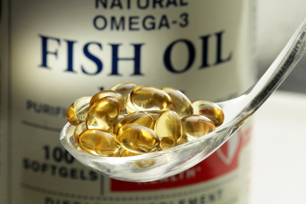 A spoonful of golden yellow fish oil or Omega-3 softgel capsules in front of the supplement bottle.