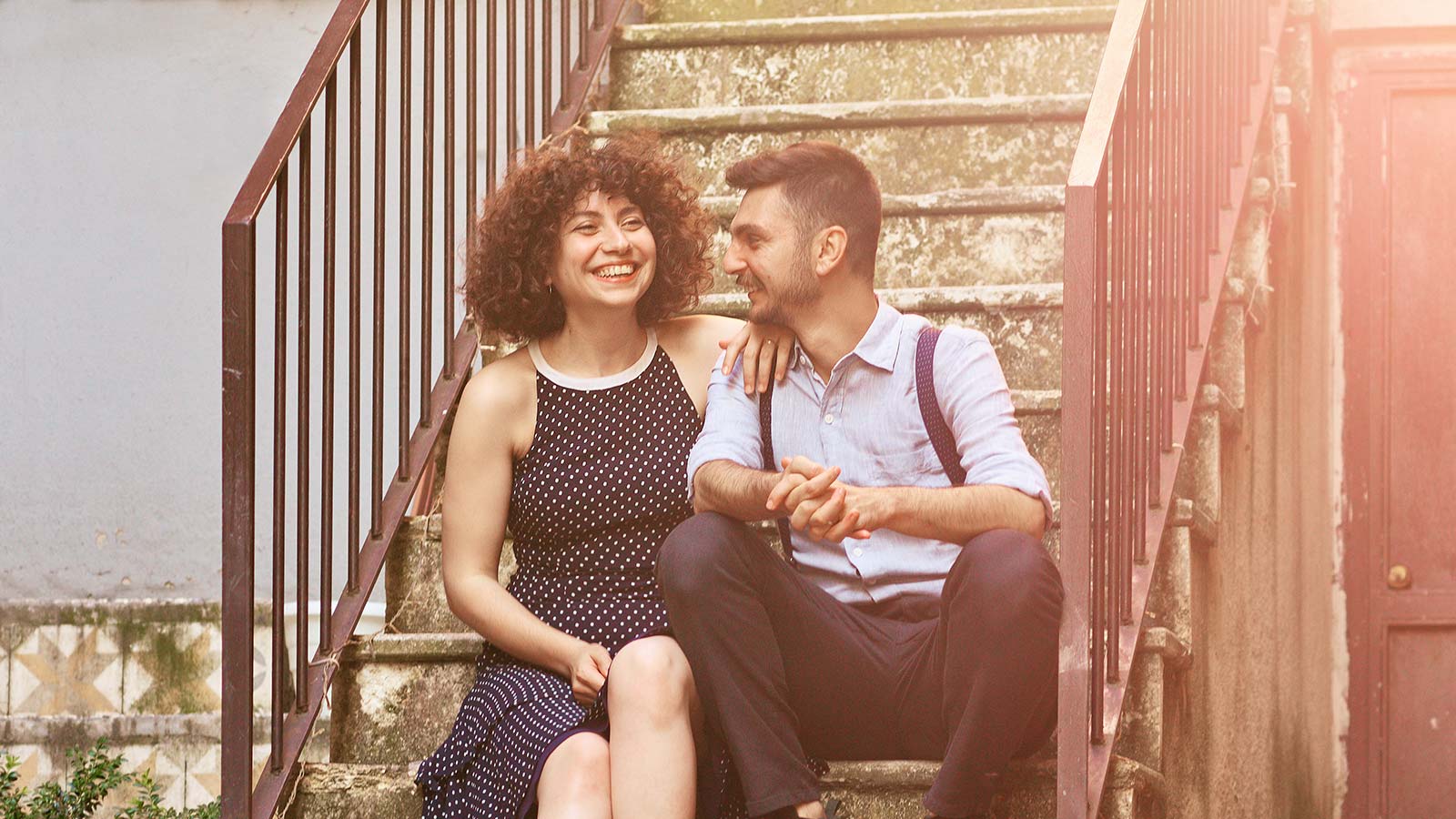 A smiling young couple is sitting together on an old stone staircase outdoors.