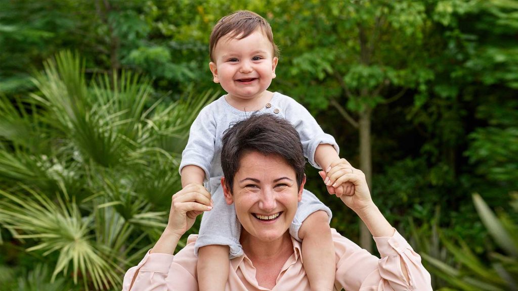 A smiling woman with short, dark hair is giving a joyous baby a shoulder ride outdoors in a lush green park or garden.