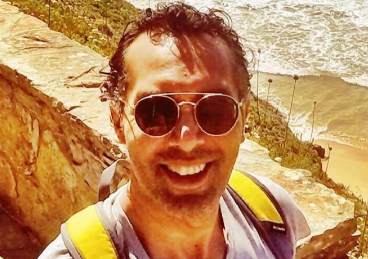 Smiling man wearing sunglasses and a backpack, standing on a coastal path with the ocean and waves visible in the background on a sunny day.