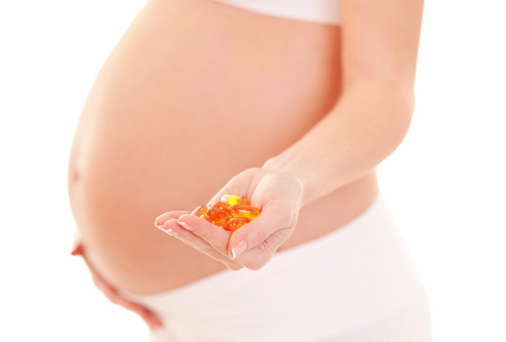Pregnant woman in white clothing holding a handful of omega-3 and multivitamin capsules, promoting prenatal nutrition and supplementation.