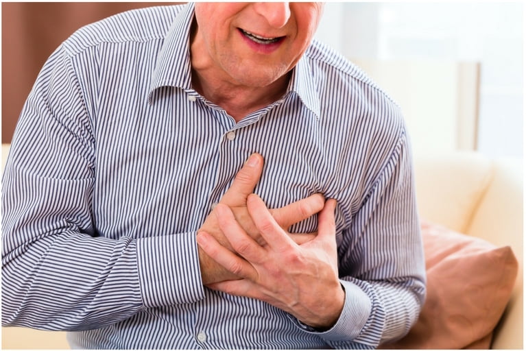 A senior man, wearing a blue and white striped button-up shirt, is sitting down and clutching his chest with both hands in an expression of discomfort or pain, indicating symptoms of a heart attack or severe chest pain.