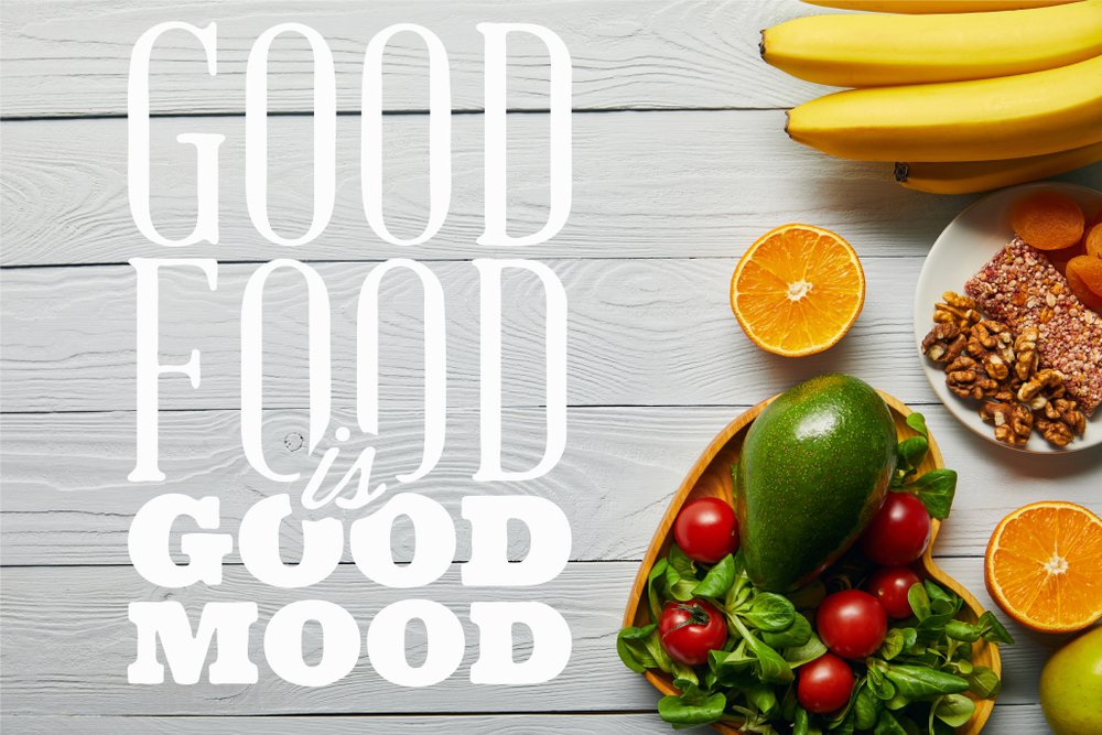 Overhead shot of healthy food (avocado, tomatoes, nuts, banana, orange slices) on a rustic wooden table with the text overlay "GOOD FOOD IS GOOD MOOD."