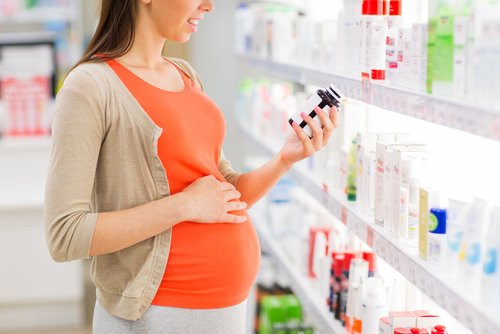 A medium shot of a pregnant woman in a pharmacy, wearing an orange top and beige cardigan, holding a bottle of medication or vitamins and reading the label.