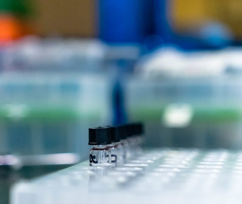 A row of small, clear glass vials with black caps sits in a white plastic test tube rack in a laboratory.