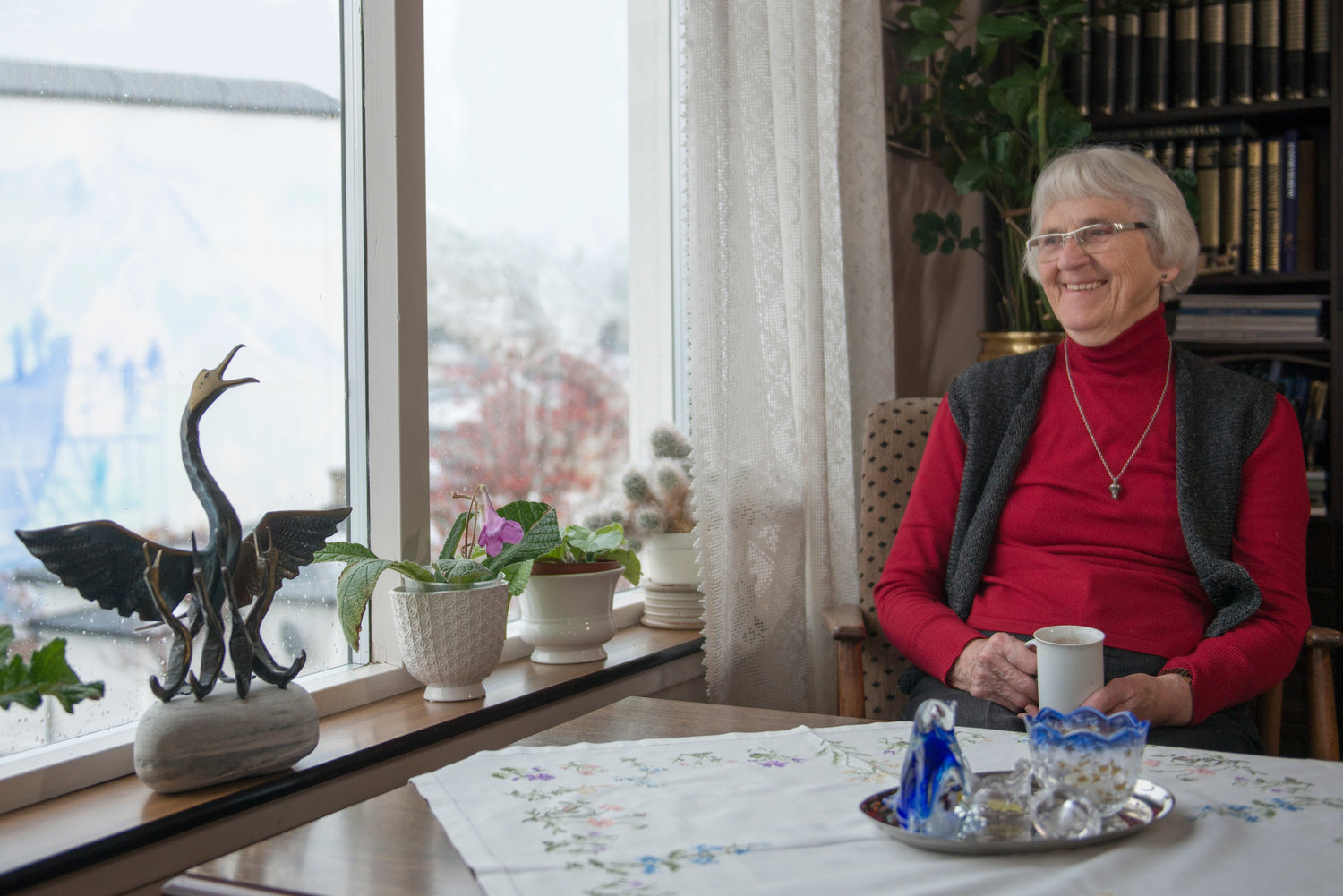 An elderly woman with short white hair and glasses is seated at a table near a large window, smiling and looking off-camera.