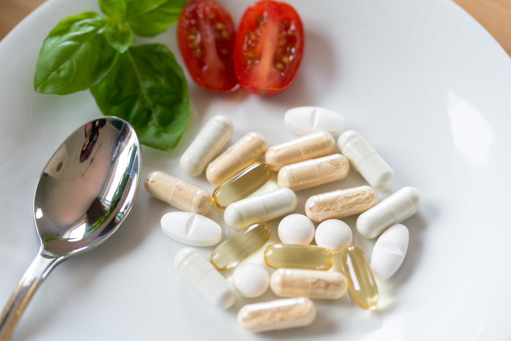 A close-up overhead shot of a white plate holds a pile of assorted vitamins and supplements, including white tablets and beige and translucent golden capsules.