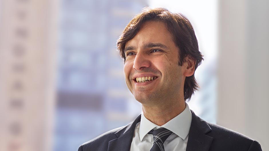 A headshot of a smiling, dark-haired man in a business suit with a white shirt and striped tie.