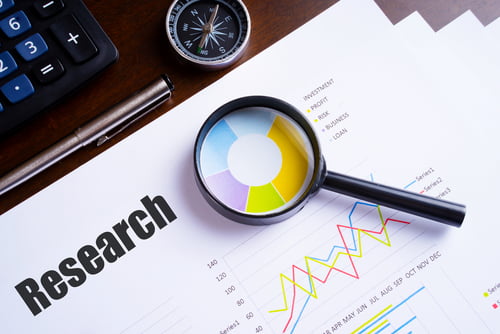 Magnifying glass placed over a colorful pie chart on a printed research report, with graphs, a compass, a pen, and a calculator on a desk.