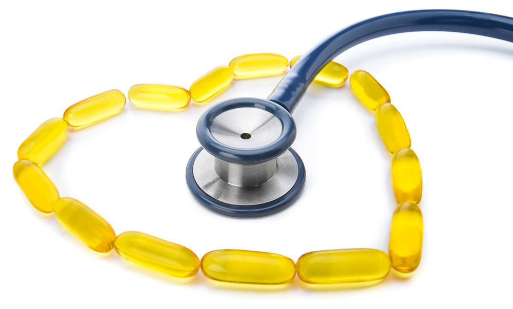 A dark blue stethoscope is resting on a white background, with its bell placed inside a heart shape outlined by yellow soft gel capsules.