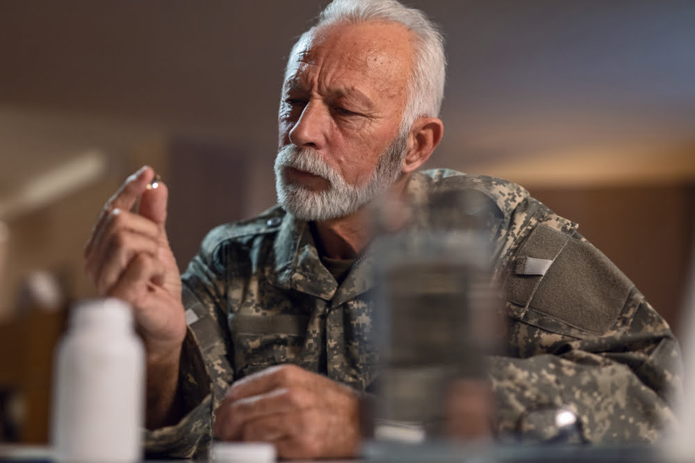 A serious-looking, older man with a gray beard and wearing a military uniform is sitting and holding up a small pill between his thumb and index finger, examining it.