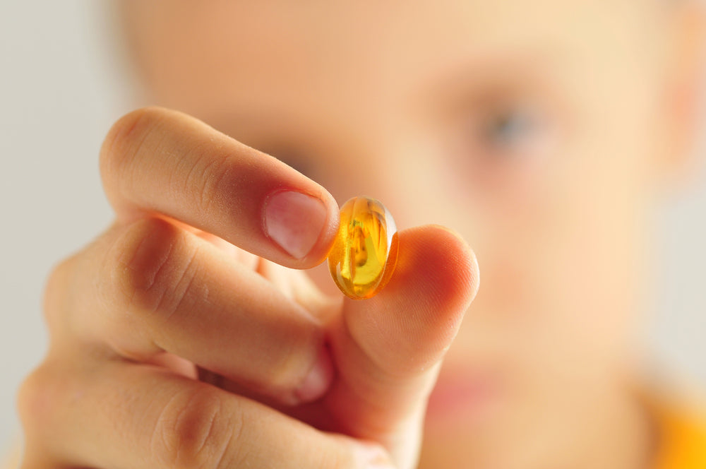 A close-up, shallow depth of field shot focuses on a single golden, translucent gel capsule being held up between a person's thumb and index finger.