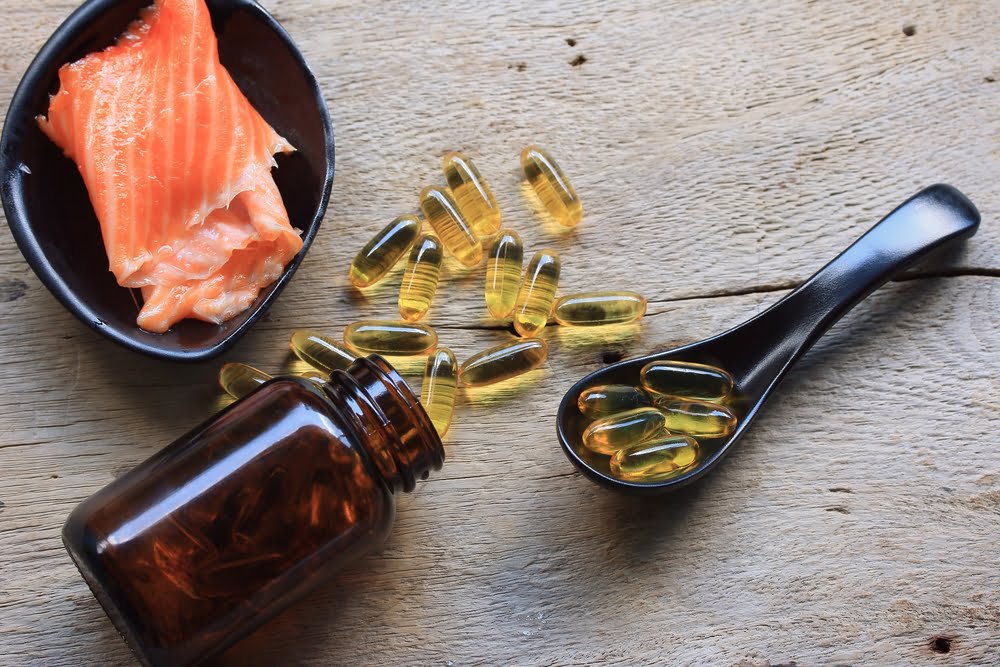 Omega-3 fish oil capsules spilled from a brown bottle next to a spoonful of capsules and a bowl containing a piece of raw salmon, placed on a rustic wooden surface.