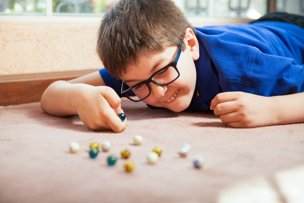 A smiling boy wearing glasses and a blue shirt is lying on a beige carpet, looking down and concentrating as he plays with a cluster of colorful marbles.