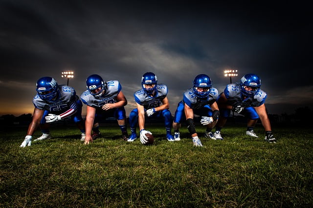 Five American football players, likely the offensive line, in uniform positioned on the line of scrimmage at night under stadium lights.