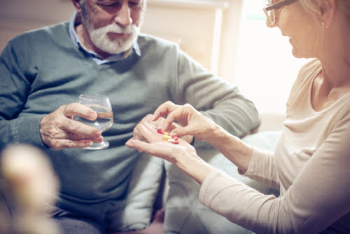 A middle-aged or elderly woman wearing glasses is offering a handful of various colored pills and capsules to an elderly man.