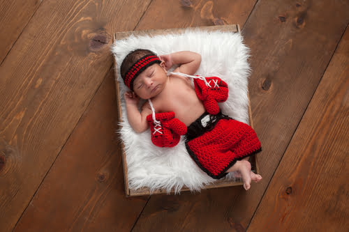 Overhead photo of a newborn baby sleeping in a wooden box while wearing a knitted boxing outfit, complete with gloves, shorts, and a headband.