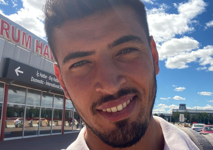 Young man with short dark hair and beard smiling in front of an airport terminal under a sunny blue sky with scattered clouds.