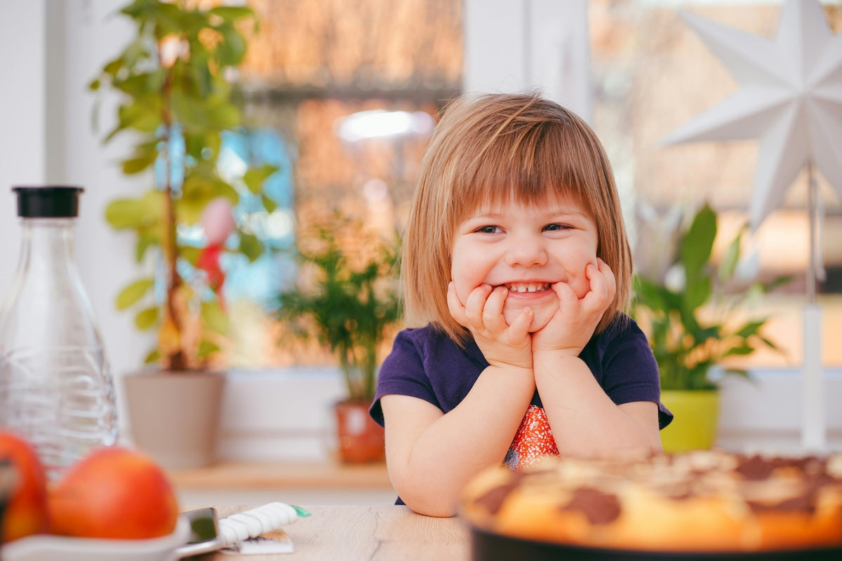 A smiling toddler with light brown hair and a fringe is sitting at a wooden table indoors, resting her chin on her hands.