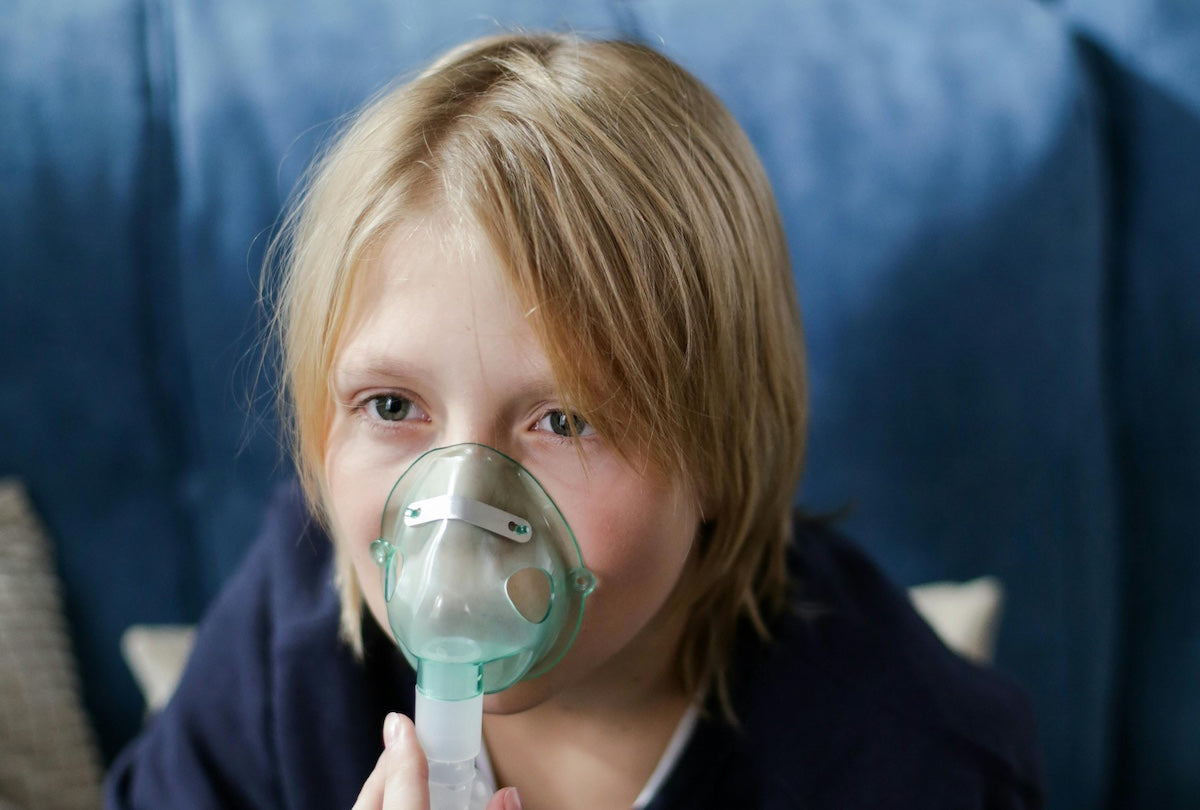 A close-up portrait of a child with blonde hair and light eyes looking directly at the camera while wearing a nebulizer mask over their nose and mouth.