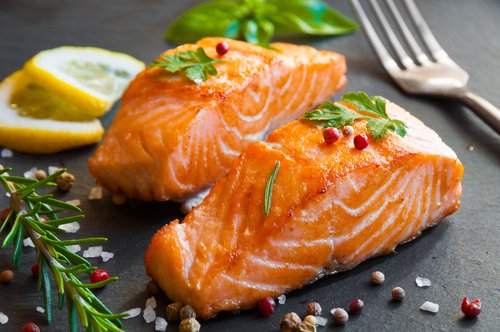 Two seasoned and cooked salmon fillets garnished with herbs, lemon slices, and peppercorns on a dark plate.