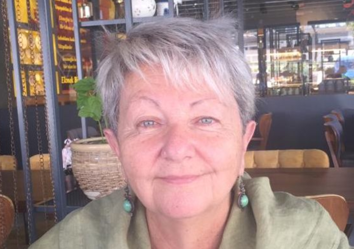 Elderly woman with short gray hair wearing green earrings and a light green top, smiling softly while seated indoors at a warmly lit restaurant.