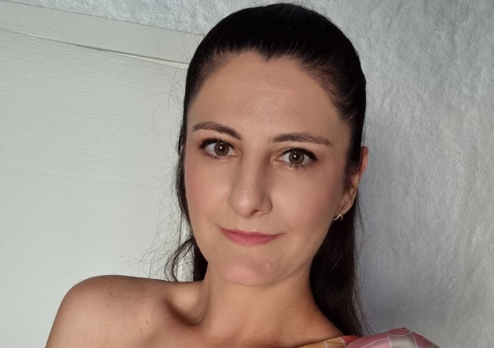 Woman with dark hair tied back, wearing light makeup and a pastel-colored top, smiling slightly while posing indoors against a textured white background.