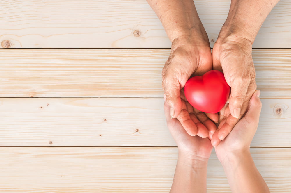 Elderly hands passing a red heart-shaped object to younger hands, symbolizing care, support, and heart health across generations.