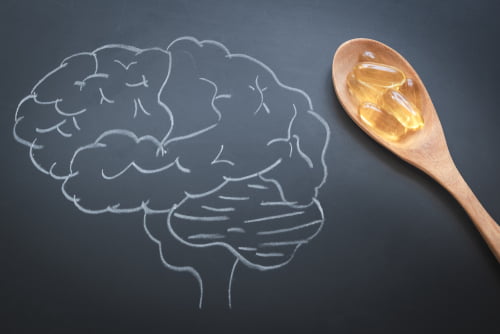 Chalk drawing of a human brain on a dark background next to a wooden spoon holding three Omega-3 capsules.