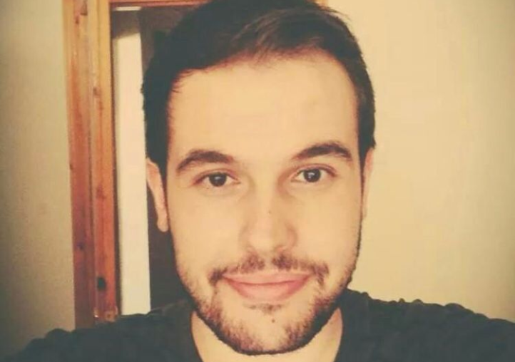 Young man with short dark hair and trimmed beard smiling slightly while taking a selfie indoors with warm lighting.