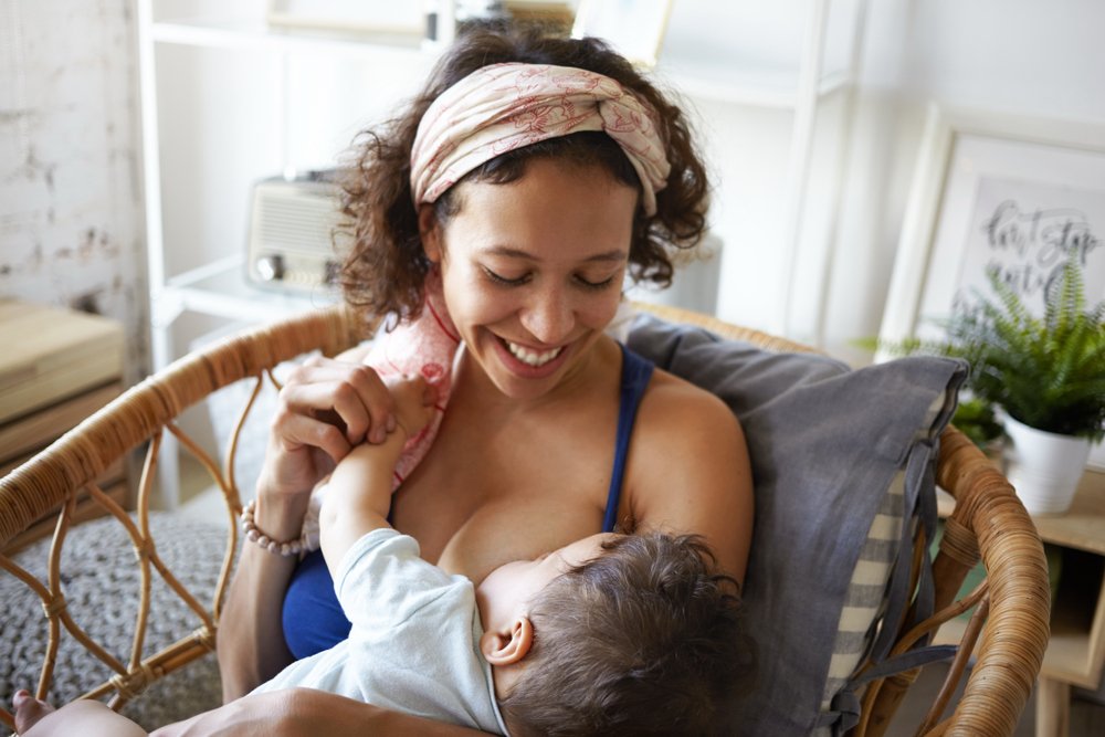 A smiling mother breastfeeding her baby while sitting in a wicker chair indoors.
